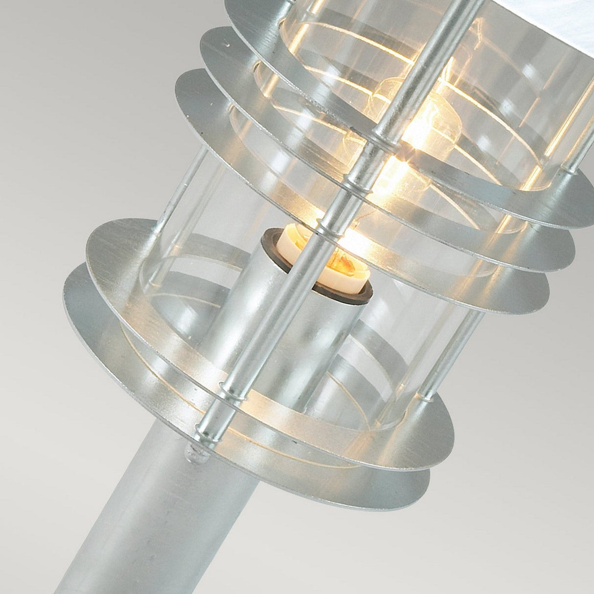 Close-up of the Stockholm 1 Light Grande Pedestal - Silver, a modern outdoor lamp post designed in Scandinavian style. It features a circular structure with transparent glass panels encasing the light bulb. The lamp is illuminated, casting a warm glow against a minimalist background.