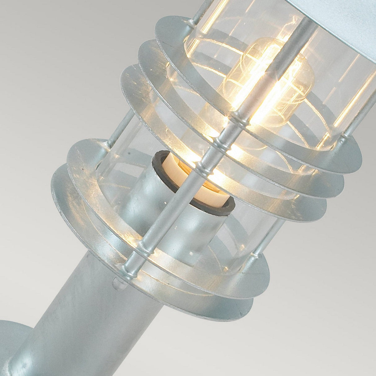 Close-up of the Stockholm 1 Light Pedestal - Silver, a modern outdoor lamp with a clear glass casing. Reminiscent of Scandinavian design, it features circular metal guard rings around the glowing bulb, set against a light gray background.