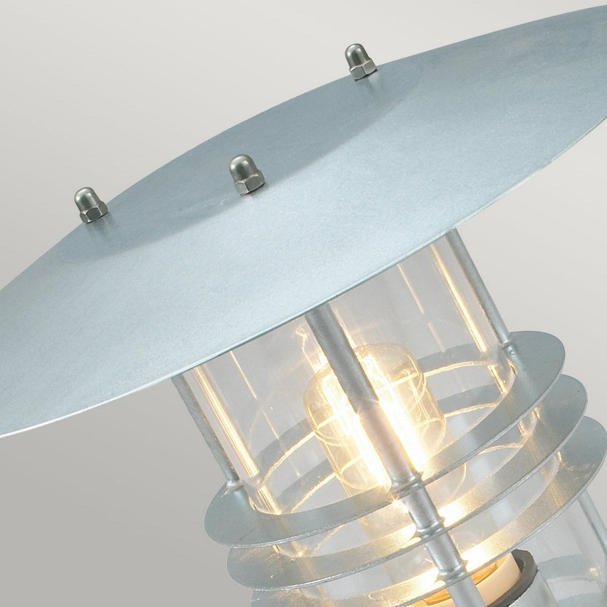 Close-up of the Stockholm 1 Light Pedestal - Silver, highlighting its Scandinavian design. It features a metallic top made from galvanized steel and a glowing bulb inside a clear cylindrical casing, encircled by sleek metal rings. The neutral gradient background enhances its minimalist charm.