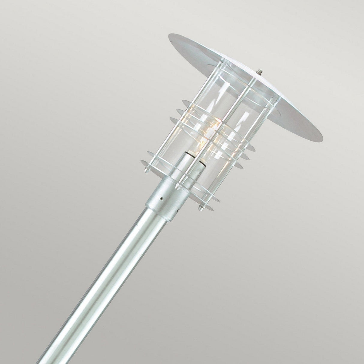 The Stockholm 1 Light Grande Lamp Post - Silver is an elegant outdoor lighting fixture with a contemporary design. It boasts a cylindrical glass casing complemented by circular metal fittings and a conical top, all made from galvanized steel. This chic lamp is mounted on a metallic pole and casts a gentle glow against a neutral backdrop.