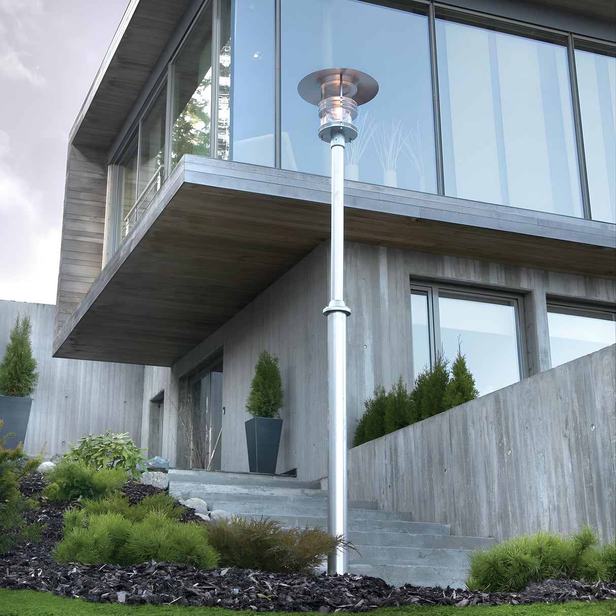 A modern house with expansive glass windows sits atop a landscaped hill. Near the building, a Stockholm 1 Light Grande Lamp Post in Silver, expertly crafted from galvanized steel, is surrounded by lush shrubs and small trees. This arrangement creates a serene and contemporary outdoor setting with chic lighting features.