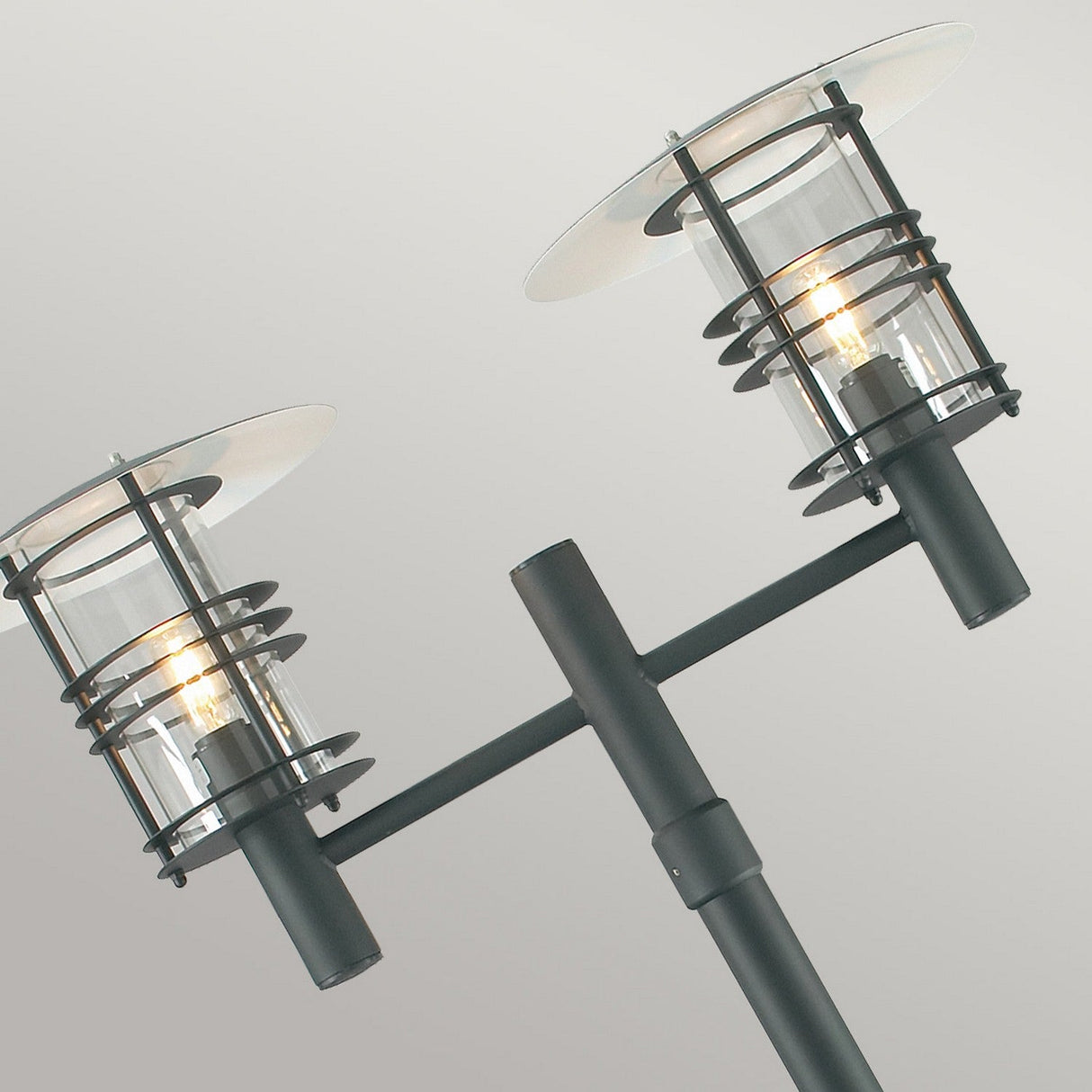 Discover the Stockholm 2 Light Twin Lamp Post - Black, a Scandinavian-inspired outdoor lighting fixture showcasing two cylindrical glass lanterns with sleek metal rings. It casts a warm, inviting glow against its black finish, seamlessly combining modern elegance with timeless charm.