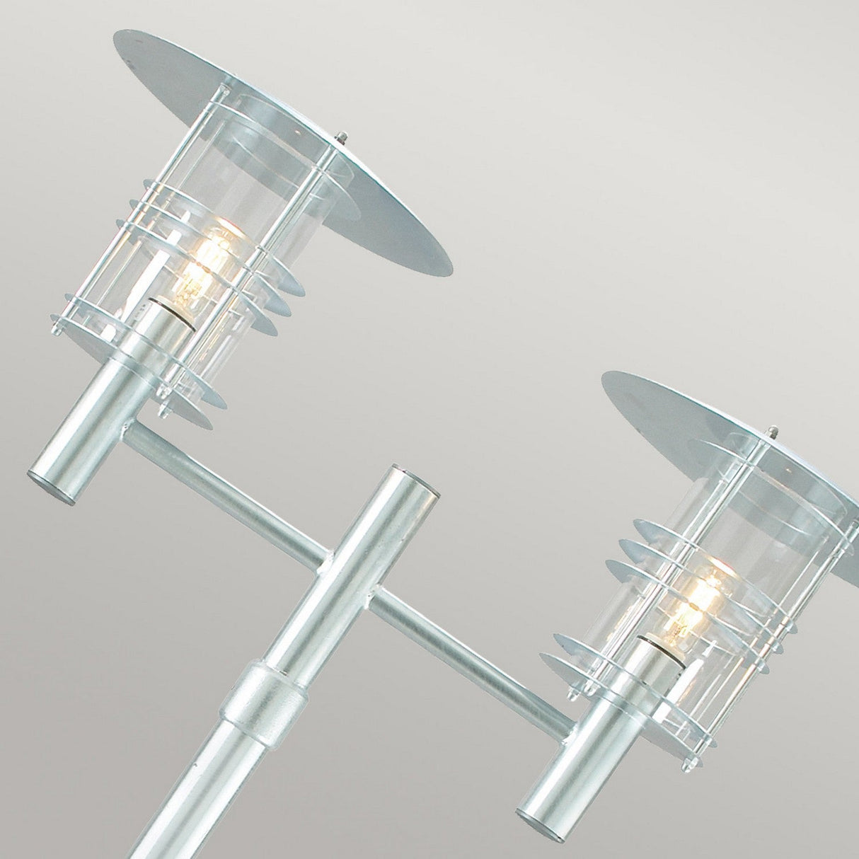 The Stockholm 2 Light Twin Lamp Post - Silver showcases a Scandinavian-inspired design with two cylindrical glass-covered bulbs on a galvanized steel pole, each adorned with circular metal caps and presented against a simple light grey backdrop.