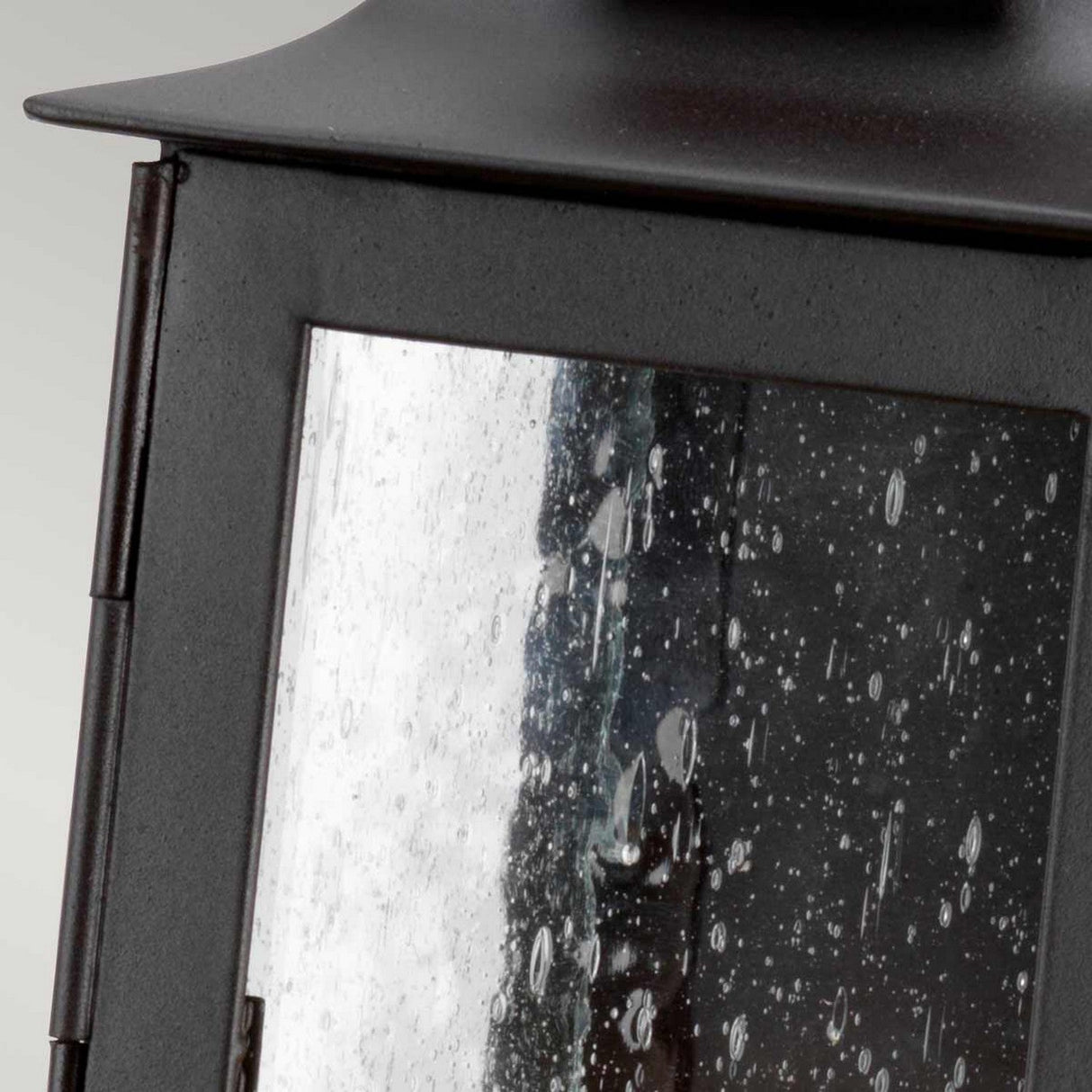 A close-up of the Stow 1 Light Outdoor Wall Light - Old Bronze showcases its black metal lantern with rain-dotted glass panes that beautifully reflect light. The droplets add a textured, antique charm to the glass, set against a background of soft, muted gray reminiscent of old bronze hues.