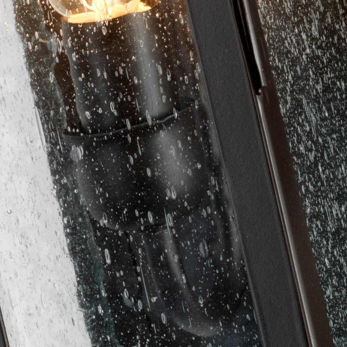 Close-up of a rain-soaked window with a blurred view of the Stow 1 Light Outdoor Wall Light - Old Bronze behind it. Raindrops stream down the glass, their textured pattern adding to the antique appeal as they softly veil the old bronze glow.