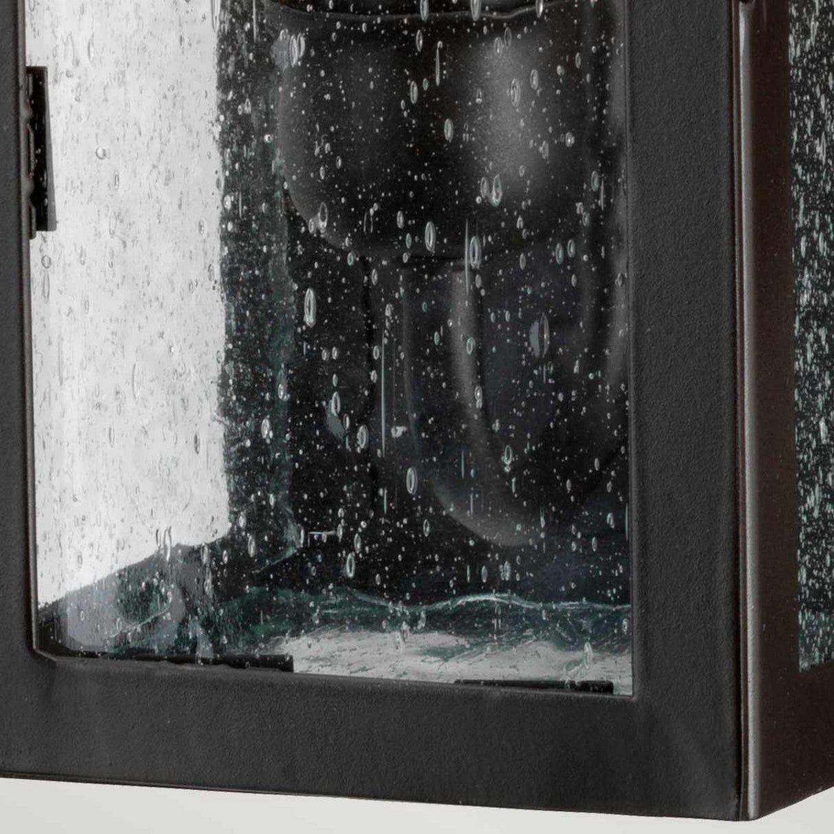 Close-up of the Stow 1 Light Outdoor Wall Light in Old Bronze, featuring a black metal frame and glass design adorned with raindrops. The lantern, exuding antique appeal, is slightly ajar to reveal its interior fixture. Set against a neutral backdrop, this piece showcases its timeless charm as an outdoor wall lighting option.