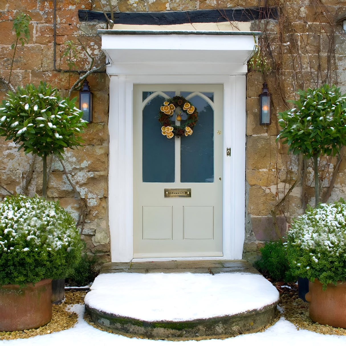 A charming cottage door features a festive wreath and is flanked by two tall potted plants lightly dusted with snow. The cozy, wintry atmosphere is heightened by the stone wall background and snow-covered steps. On either side of the door, an antique appeal is enhanced by two Stow 1 Light Outdoor Wall Lights in Old Bronze adorning the wall.