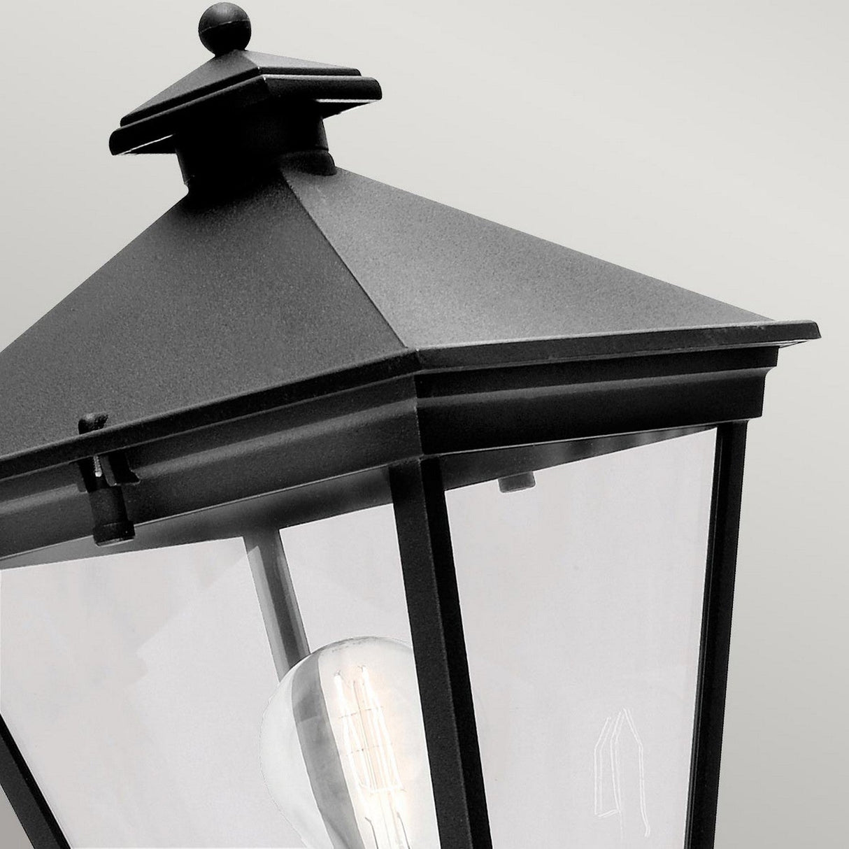 Close-up of the Turin Outdoor 1 Light Up Wall Lantern - Black, showcasing its simple geometric design crafted from durable die-cast aluminum. It features a clear glass panel revealing a visible light bulb inside and boasts an IP54 rating. The lantern is mounted against a light gray background.
