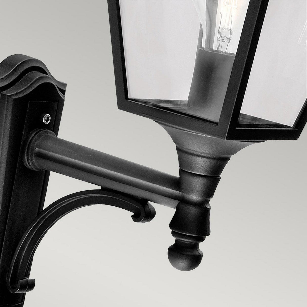 Close-up of the Turin Outdoor 1 Light Up Wall Lantern in black, made from durable die-cast aluminum with a clear glass shade, showcasing a visible light bulb inside. The lantern, which features an IP54 rating, is attached to a decorative mounting bracket against a neutral background.