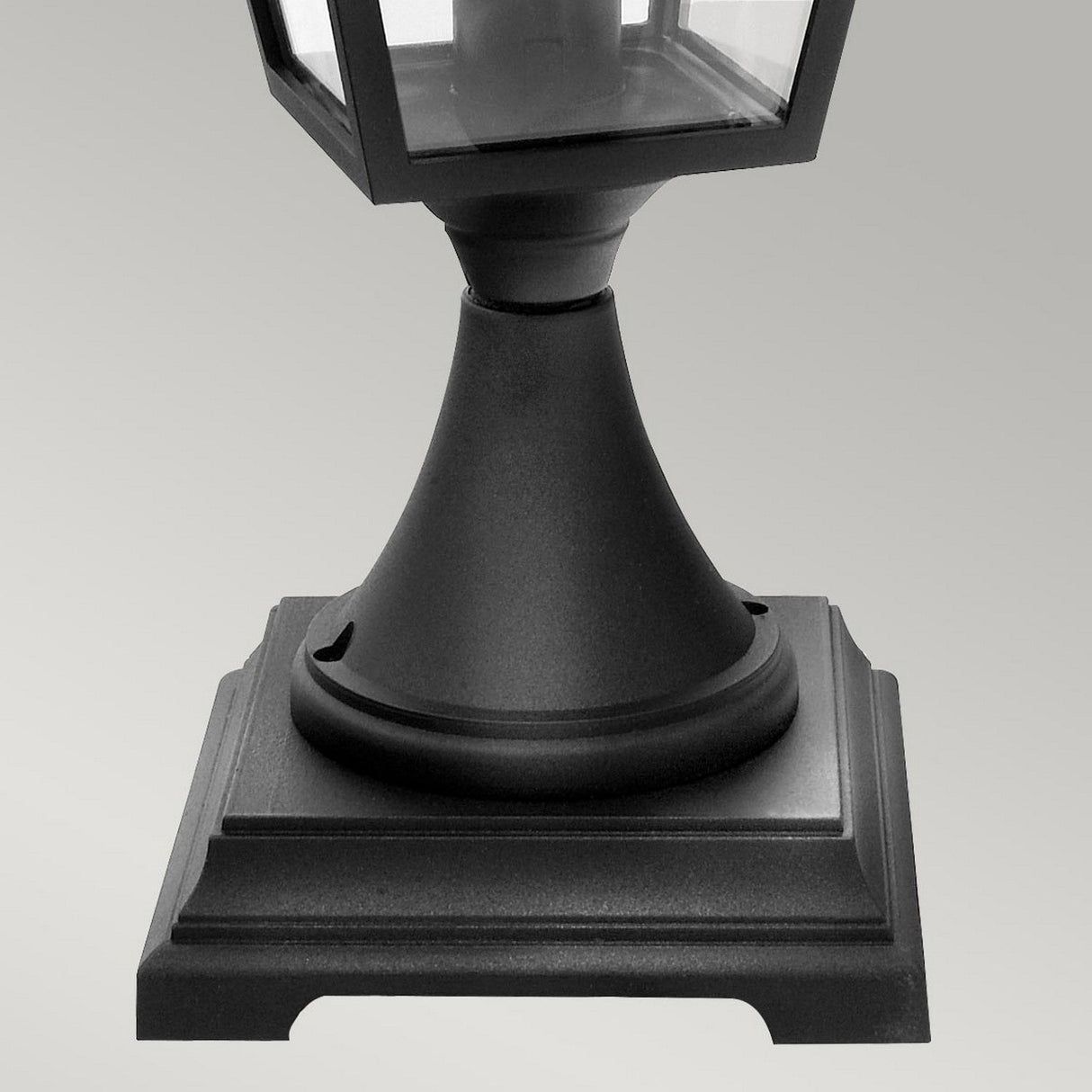 Close-up of the base of the Turin 1 Light Pedestal - Black, an outdoor lantern made from die-cast aluminum with a square, stepped design. It is mounted on a tapered stand, exuding a minimalist and modern aesthetic. The lantern is suitable for various weather conditions due to its IP54 rating, set against a plain gray background.
