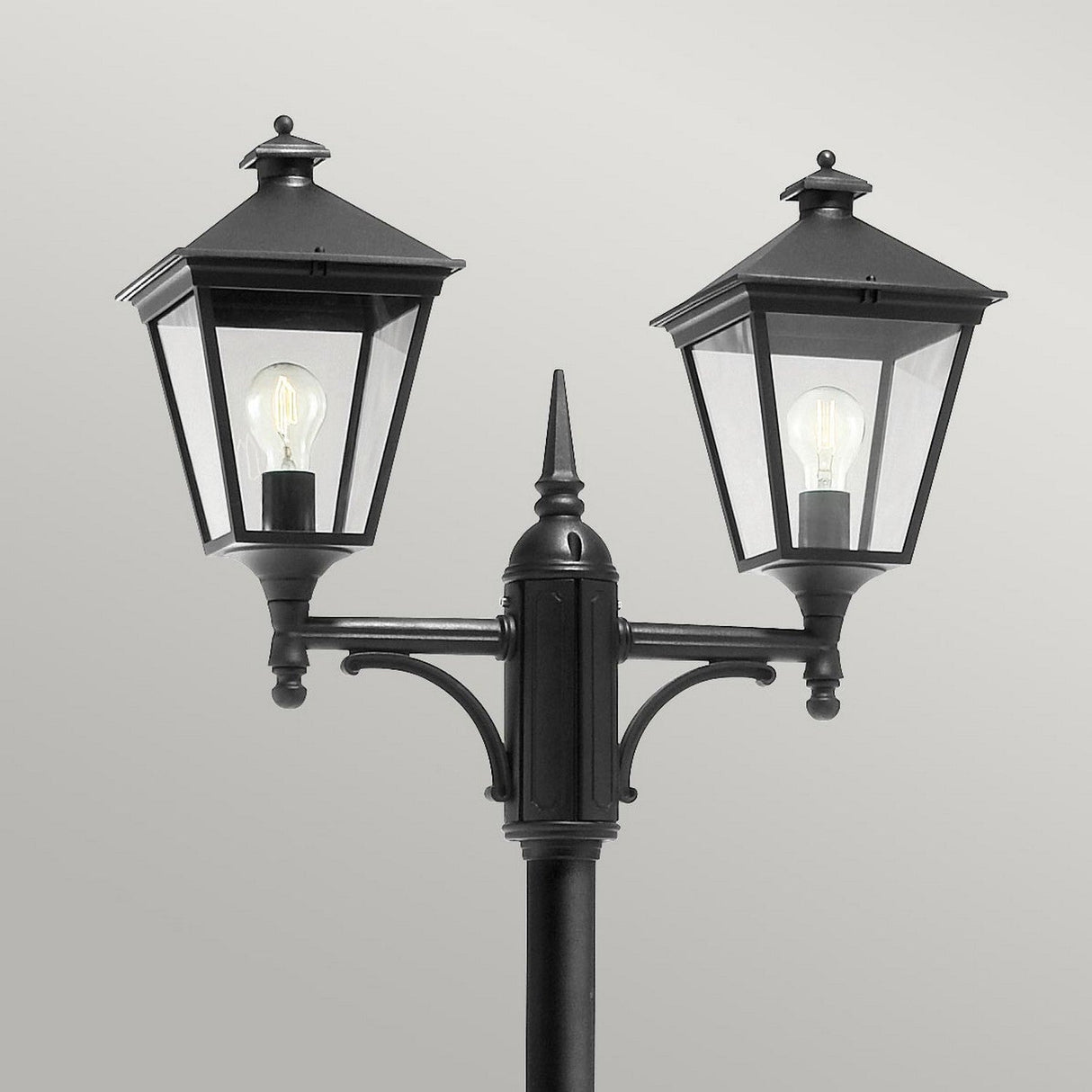 The Turin 2 Light Twin Lamp Post - Black features a black metal structure crafted from die-cast aluminum with clear glass panels that showcase visible light bulbs. It stands against a plain gray background, offering an IP54 rating and a classic angular design reminiscent of a vintage style.