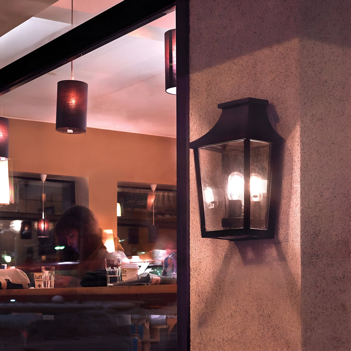 The Turin Outdoor 1 Light Half Wall Lantern - Black, crafted from die-cast aluminium and featuring an IP54 rating, is mounted on an exterior wall beside a large window. Inside, a person sits at a table in a cozy cafe with hanging lights and paper cups scattered about, beautifully captured during nighttime.