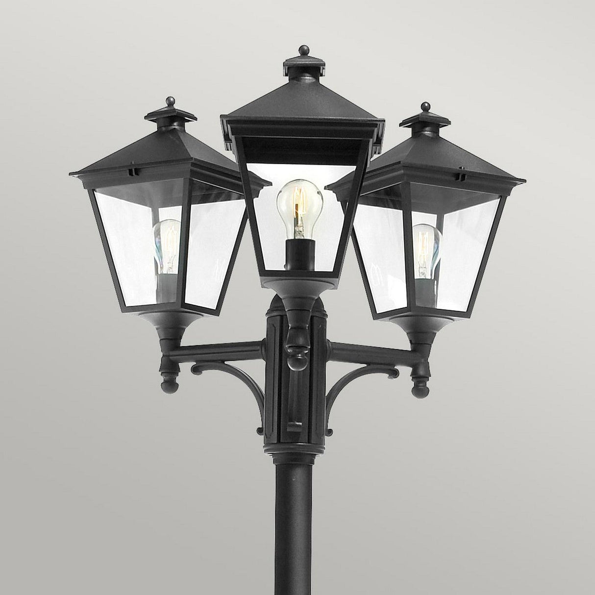 The Turin 3 Light Triple Lamp Post - Black is an ideal choice for garden illumination, featuring three lantern-style lamps with clear glass panels mounted on a single post. The central lamp stands slightly taller, set against a plain gray background to enhance the outdoor lighting appeal.