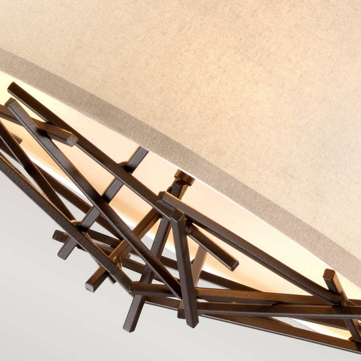 Close-up of a modern, geometric ceiling light fixture featuring a beige lampshade and decorative metal framework composed of intersecting lines that resemble twigs or branches. This sculptural element, known as the Taiko 3 Light Chandelier - Olde Bronze, softly illuminates the surrounding area.