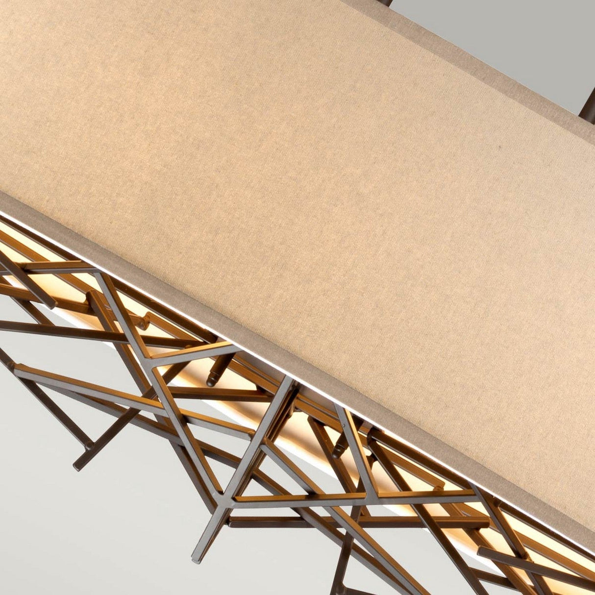 Close-up of the Taiko 5 Light Island Suspension Light - Olde Bronze, showcasing its rectangular beige shade enveloped in an angular crisscrossing metal design. When turned on, it casts a warm glow.