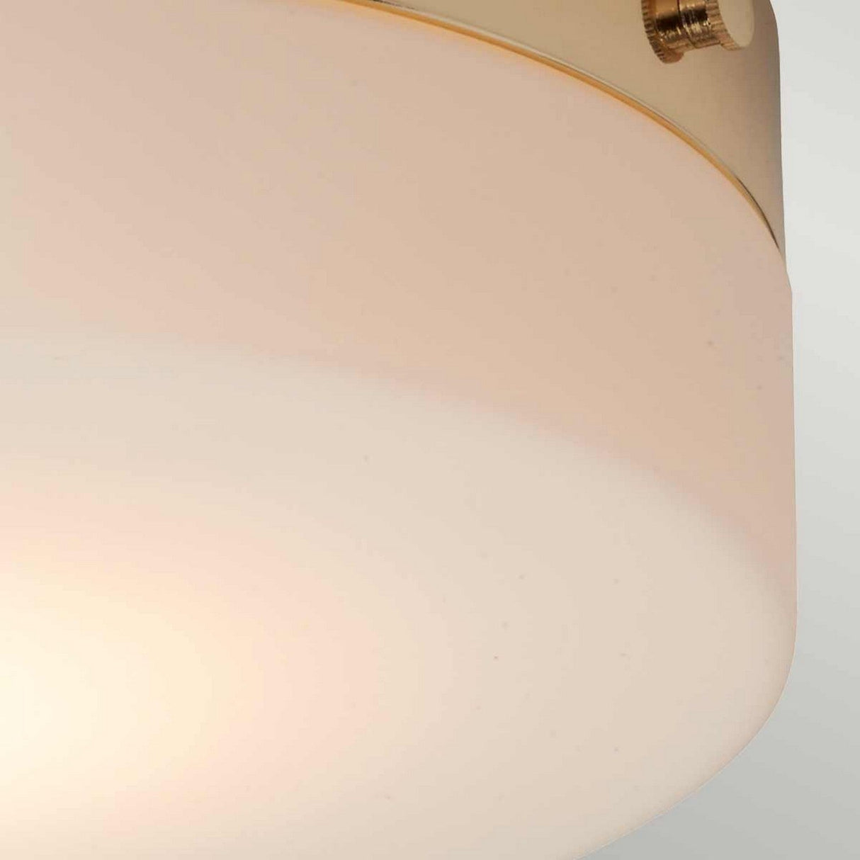 A close-up of the Tamar 1 Light Large Flush in polished gold, featuring a round, cream-colored design with a smooth, matte finish, is showcased. This energy-efficient lighting solution is attached by a sleek, metallic-colored band with visible screws. The softly lit background highlights the fixture's minimalist design.