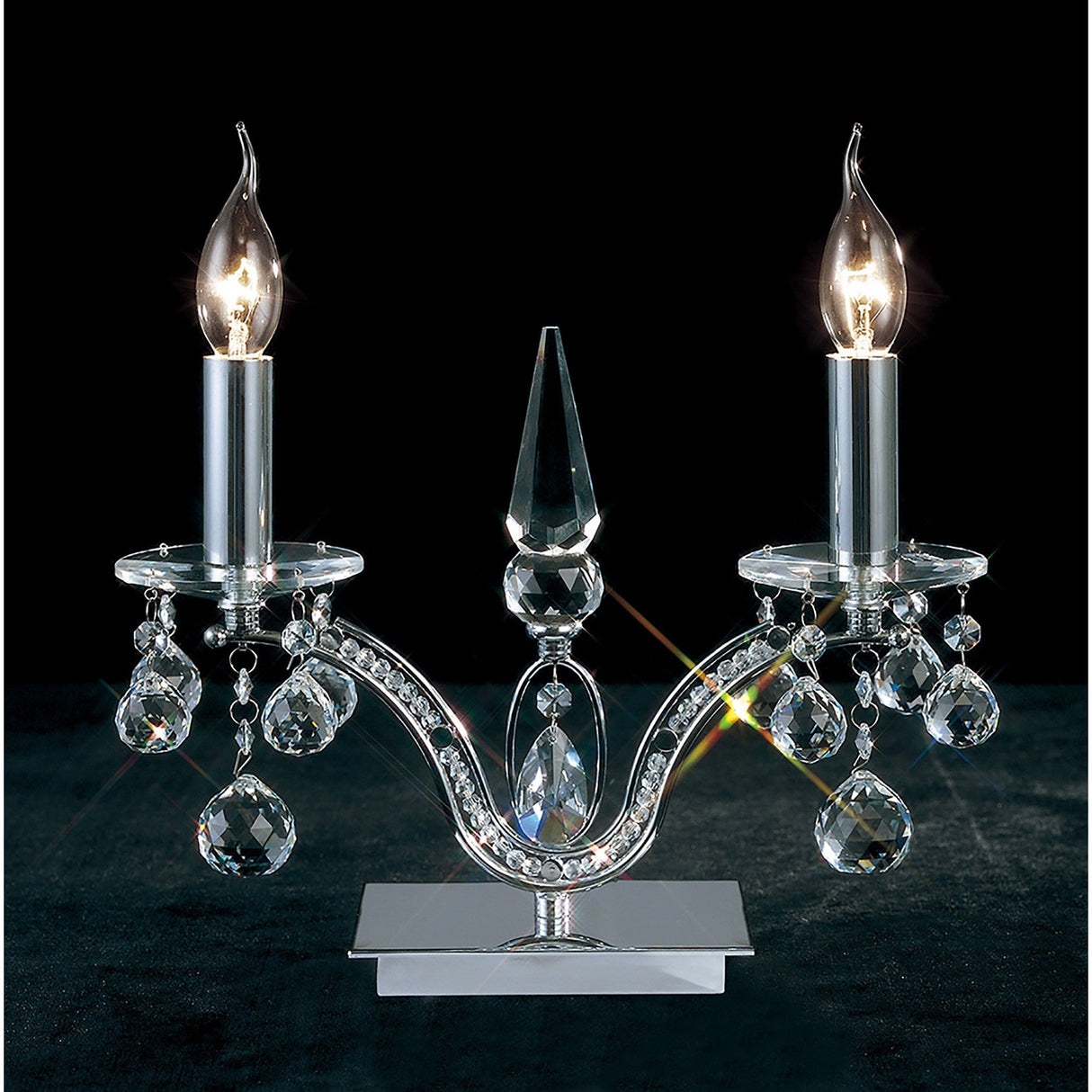 The Tara 2 Light Crystal Table Lamp in polished chrome features two candle-shaped bulbs, crystal accents, and a faceted centerpiece, creating an elegant shimmer reminiscent of classic charm against a dark backdrop.