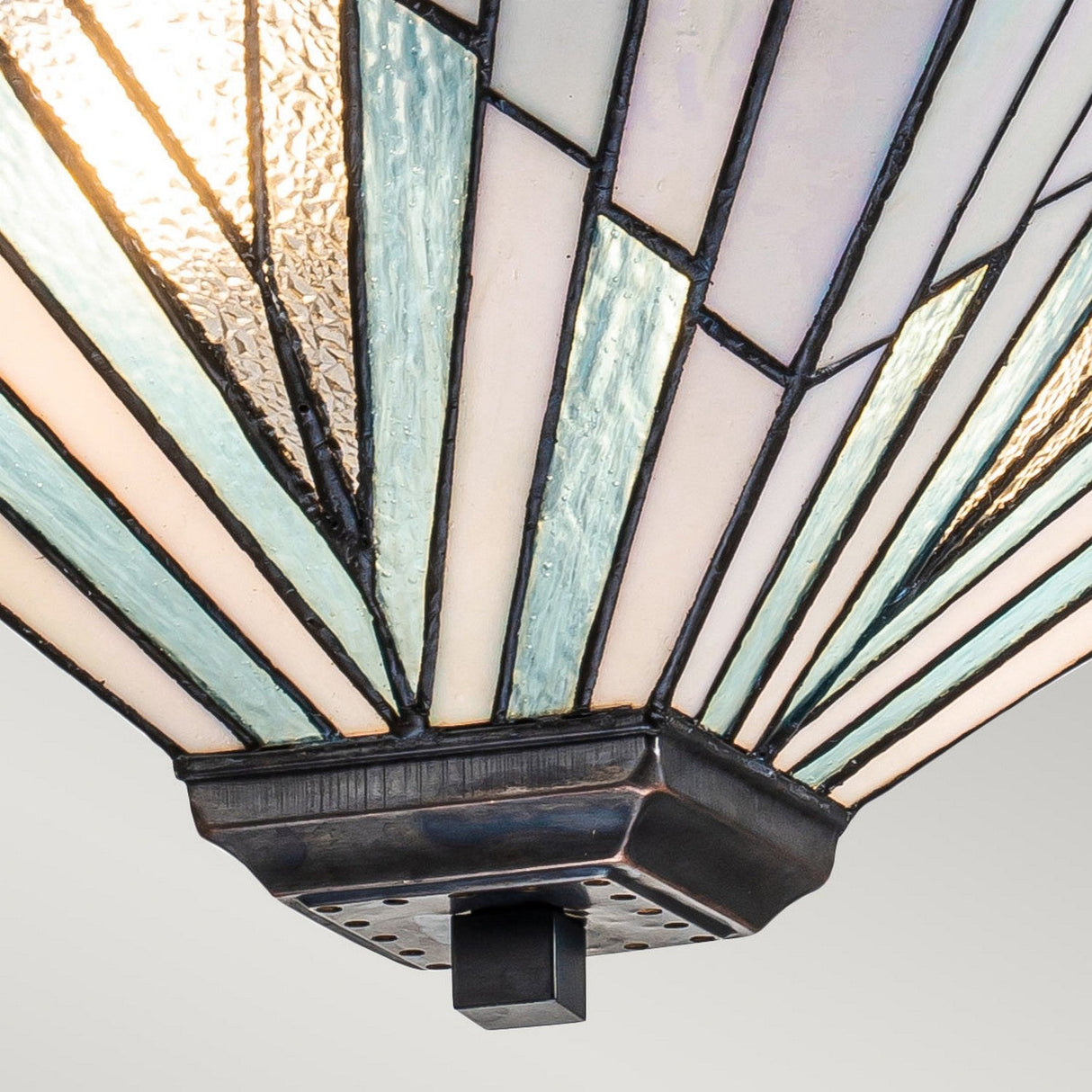 A close-up view of the Alderley 2 Light Trio Mount Tiffany Pendant showcases a stained glass lampshade with geometric patterns in blue, white, and clear glass segments, forming a fan-like shape reminiscent of an Art Deco style. This exquisite design is mounted on a vintage bronze square base.
