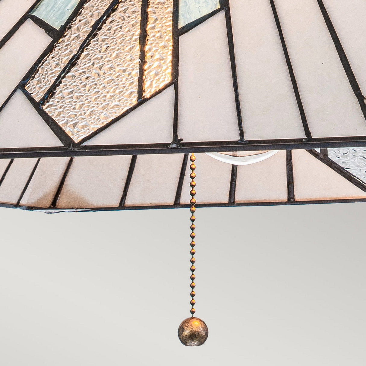 A close-up of the Alderley 2 Light Tiffany Pendant in Vintage Bronze features a stained glass lampshade with Art Deco styling, displaying a geometric pattern in white, gray, and textured glass. A vintage bronze pull chain with a round bead hangs from the lamp, set against a plain gray background.