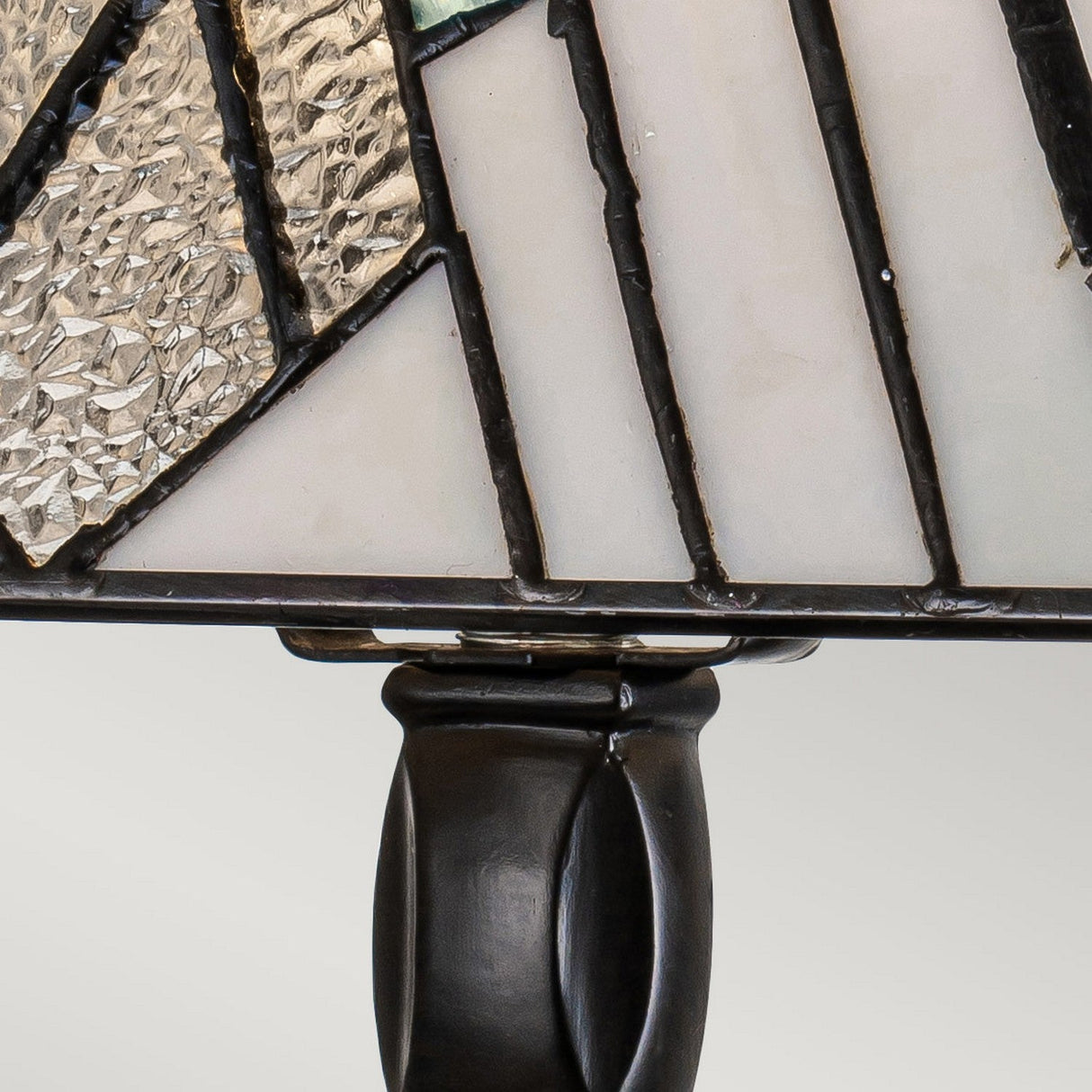 Close-up of the Alderley 1 Light Medium Table Lamp in a Tiffany-style design, showcasing abstract geometric patterns. This vintage bronze table lamp combines textured and smooth glass pieces in clear, white, and green hues, all bordered by dark metal strips and supported by a sleek, curved stand.