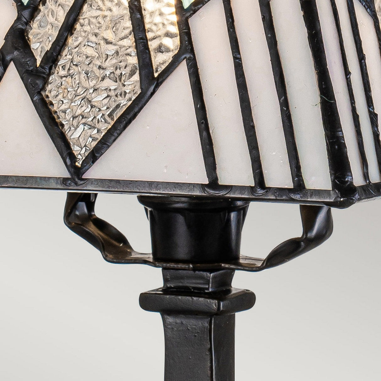 Close-up of the Alderley 1 Light Small Table Lamp, showcasing a Tiffany-style lamp shade with a geometric design. The glass panels are bordered by black metal strips and feature both textured and smooth frosted sections. Its vintage bronze base highlights intricate metalwork, creating a standout piece of decorative lighting.