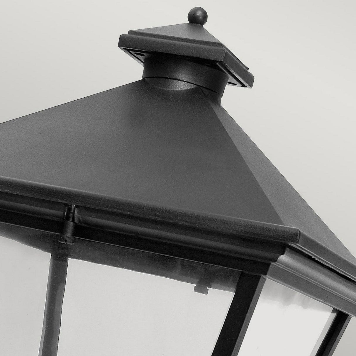 Close-up view of the top portion of the Turin Large Outdoor 1 Light Up Wall Lantern in black, crafted from die-cast aluminum with a pyramidal metal roof and glass panels. Boasting an IP54 rating, this lantern seamlessly combines durability and elegance against a neutral gray backdrop.