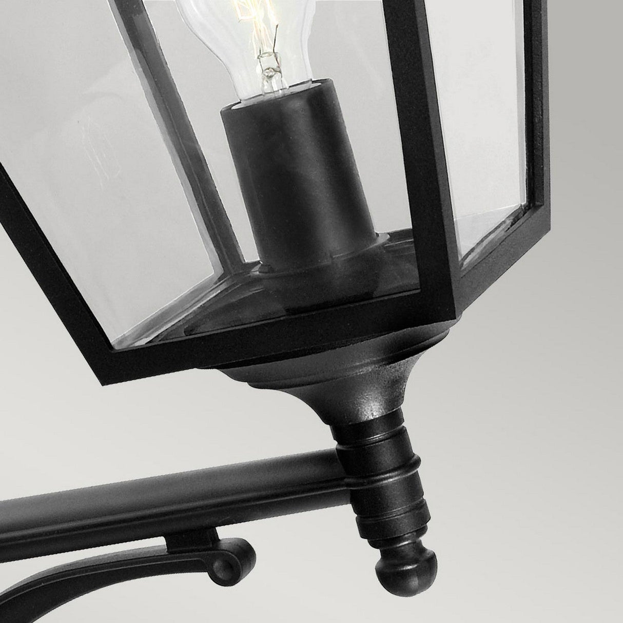 Close-up of the Turin Large Outdoor 1 Light Up Wall Lantern - Black, made from die-cast aluminium and featuring clear glass panes that showcase a visible light bulb. The design boasts sleek, angular lines with a modern aesthetic against a plain gray background and has an IP54 rating for weather resistance.