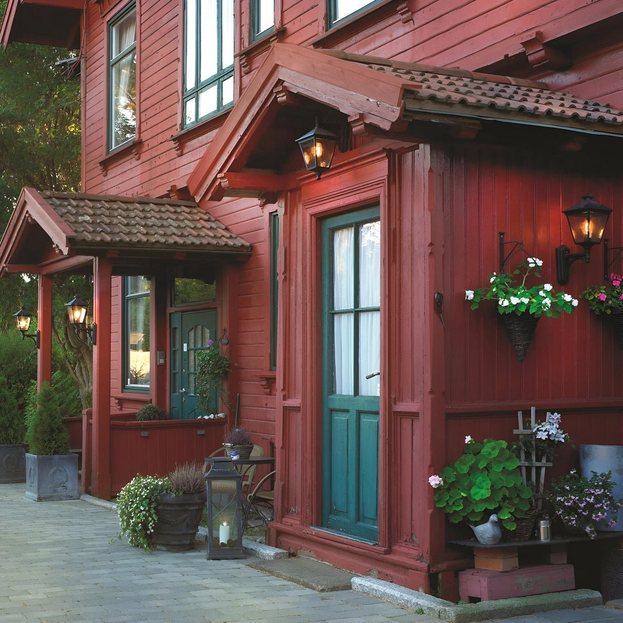 A charming red wooden building with two green doors features Turin Large Outdoor 1 Light Up Wall Lanterns in a sleek black finish, boasting an IP54 rating. Potted plants adorn the entrance, while small awnings and windows with white curtains add character. A paved path leads past this die-cast aluminium-accented structure, surrounded by greenery.
