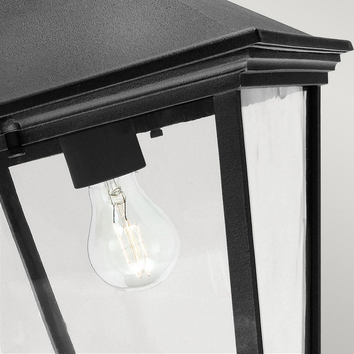 A close-up view of the Turin Large Outdoor 1 Light Down Wall Lantern in black showcases its angular design with clear glass panels and a powder-coated finish, revealing an exposed incandescent light bulb inside. This highly weather-resistant fixture is presented against a plain gray backdrop.