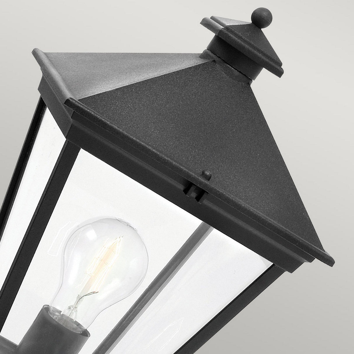 Close-up of a Turin Large 1 Light Pedestal in black metal, featuring a clear glass pane and visible light bulb inside. Made from die-cast aluminum, the lantern has a pyramid-shaped top and is set against a neutral background.