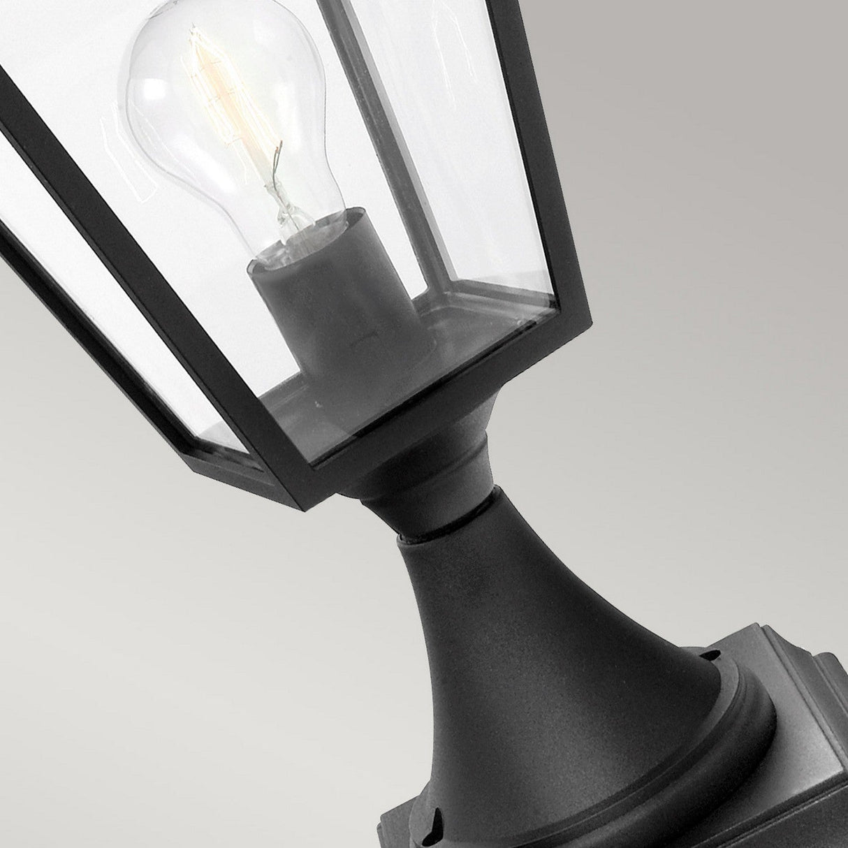 A close-up of a black outdoor lamp, expertly crafted from die-cast aluminum and featuring a clear glass shade with a visible light bulb inside. This striking piece, the Turin Large 1 Light Pedestal - Black, is displayed against a neutral grey background.
