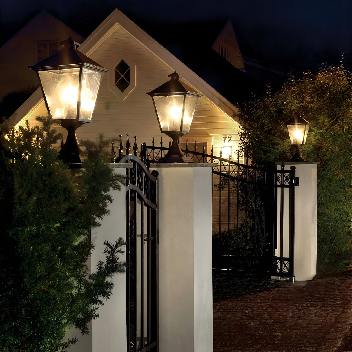 In a nighttime scene, a well-lit pathway leads to a house, illuminated by elegant Turin Large 1 Light Pedestal - Black lantern-style lights made of die-cast aluminium. The entrance is framed by a black wrought iron gate and stone pillars, while trees and bushes line the path, showcasing exquisite outdoor lighting.