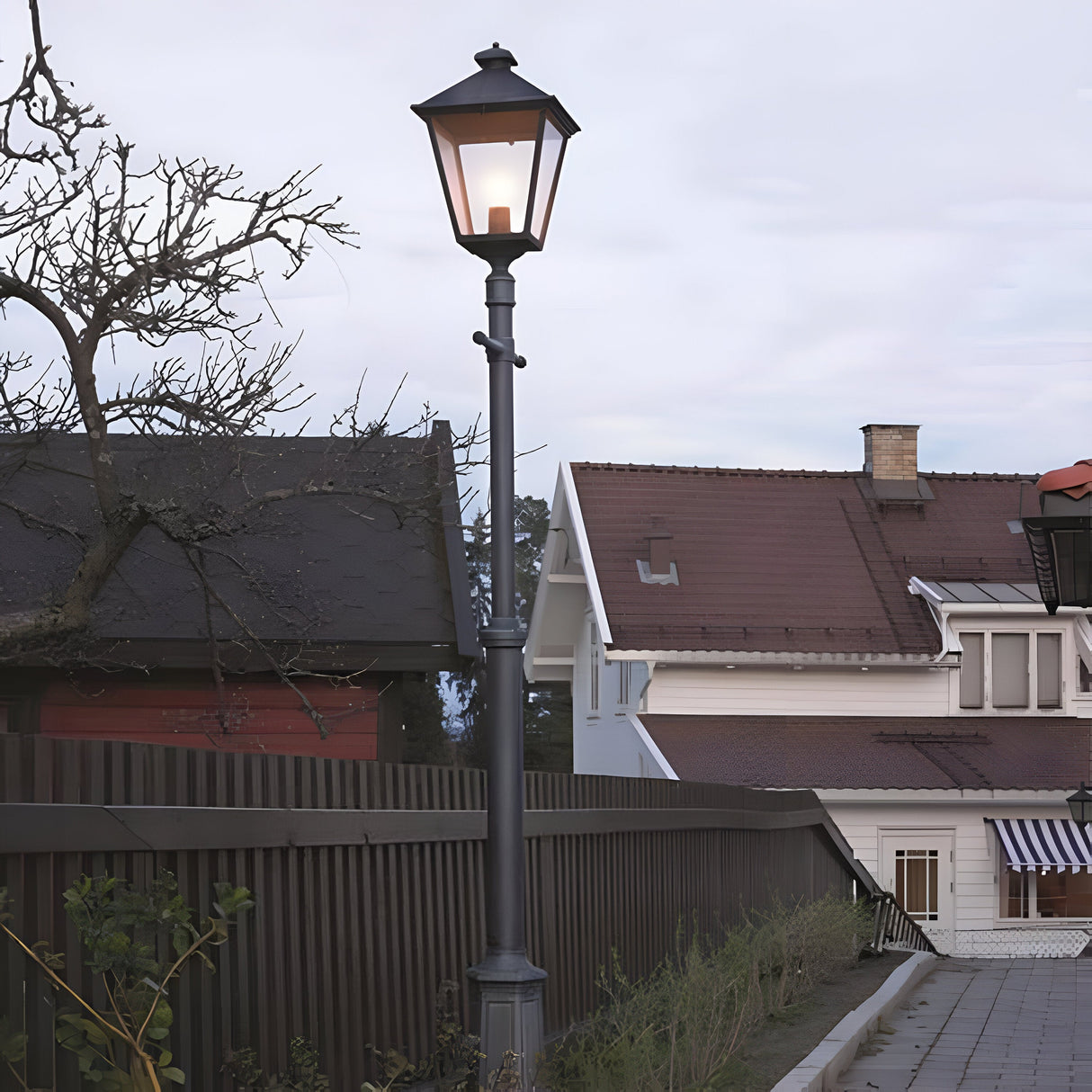 A Turin Large 1 Light Single Post in black, with its vintage-style design and die-cast aluminum construction, casts a warm glow over a tranquil neighborhood street framed by houses. The cloudy sky hints at an early evening atmosphere, while a wooden fence and bare tree branches enclose this IP54-rated outdoor lighting scene.