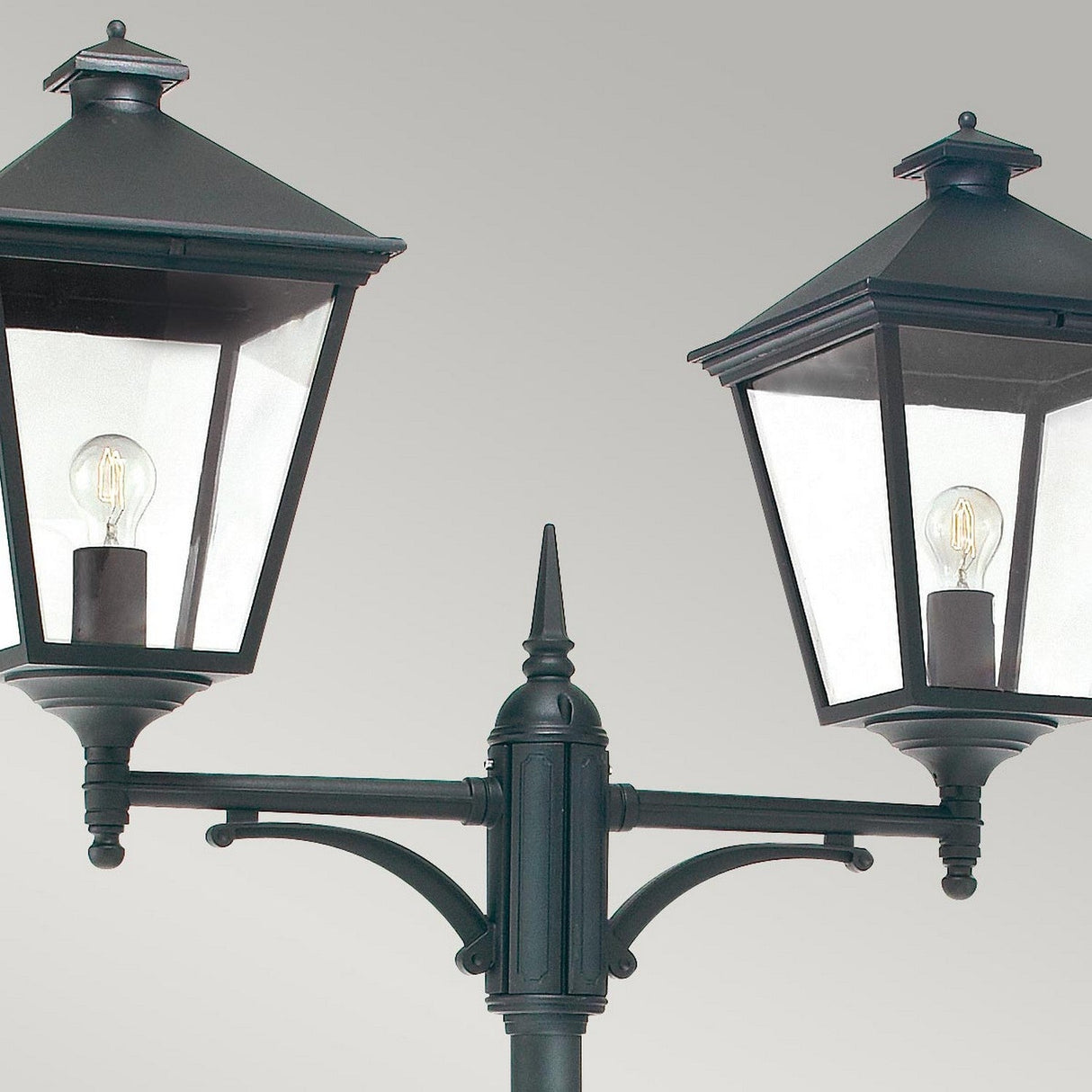 A close-up of the Turin Large 2 Light Twin Lamp Post - Black highlights elegant outdoor lighting design. Each lantern, made from durable die-cast aluminum, displays a black metal frame with clear glass panels, showcasing visible light bulbs. Mounted on one pole, it features exquisite decorative detailing.