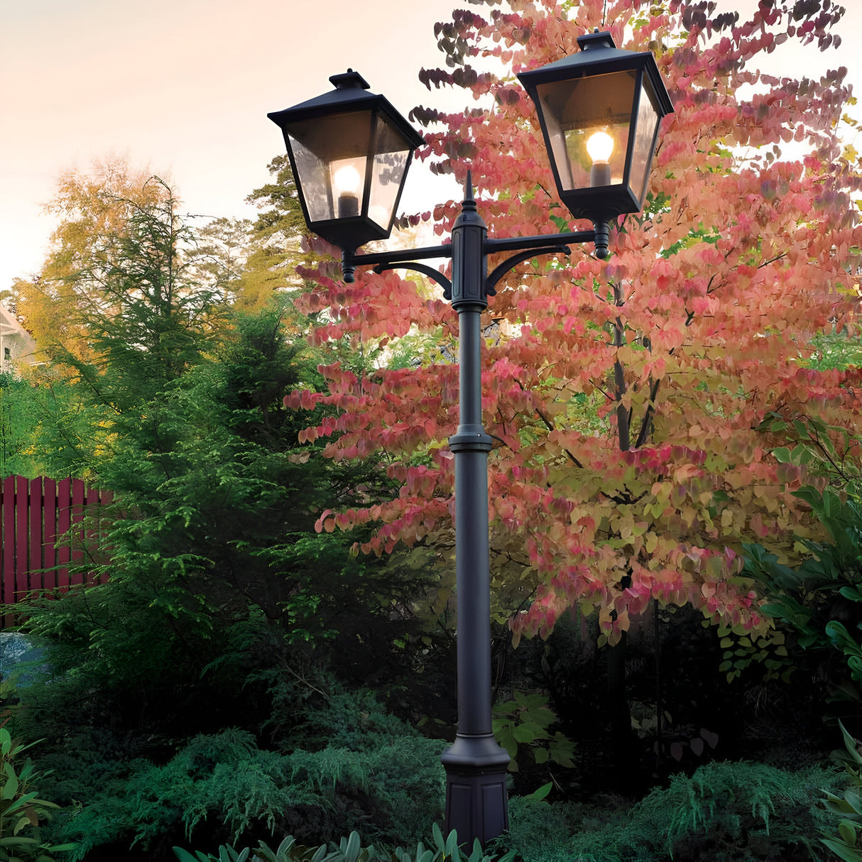 The Turin Large 2 Light Twin Lamp Post - Black, made from robust die-cast aluminum, stands gracefully amidst vibrant autumn trees adorned with red, orange, and green leaves. This picturesque scene is beautifully highlighted against a red fence under a soft evening sky, exemplifying sophisticated outdoor illumination.