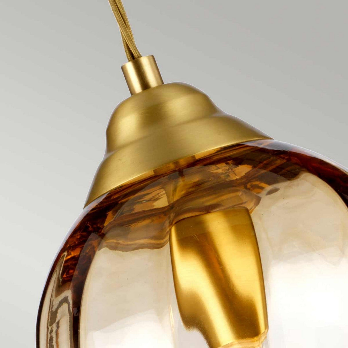 Close-up of the Tiber 1 Light Pendant, showcasing its golden metallic fixture and amber glass shade suspended from a cord. The brushed brass finish shines against the gray background, enhancing elegance in any space.