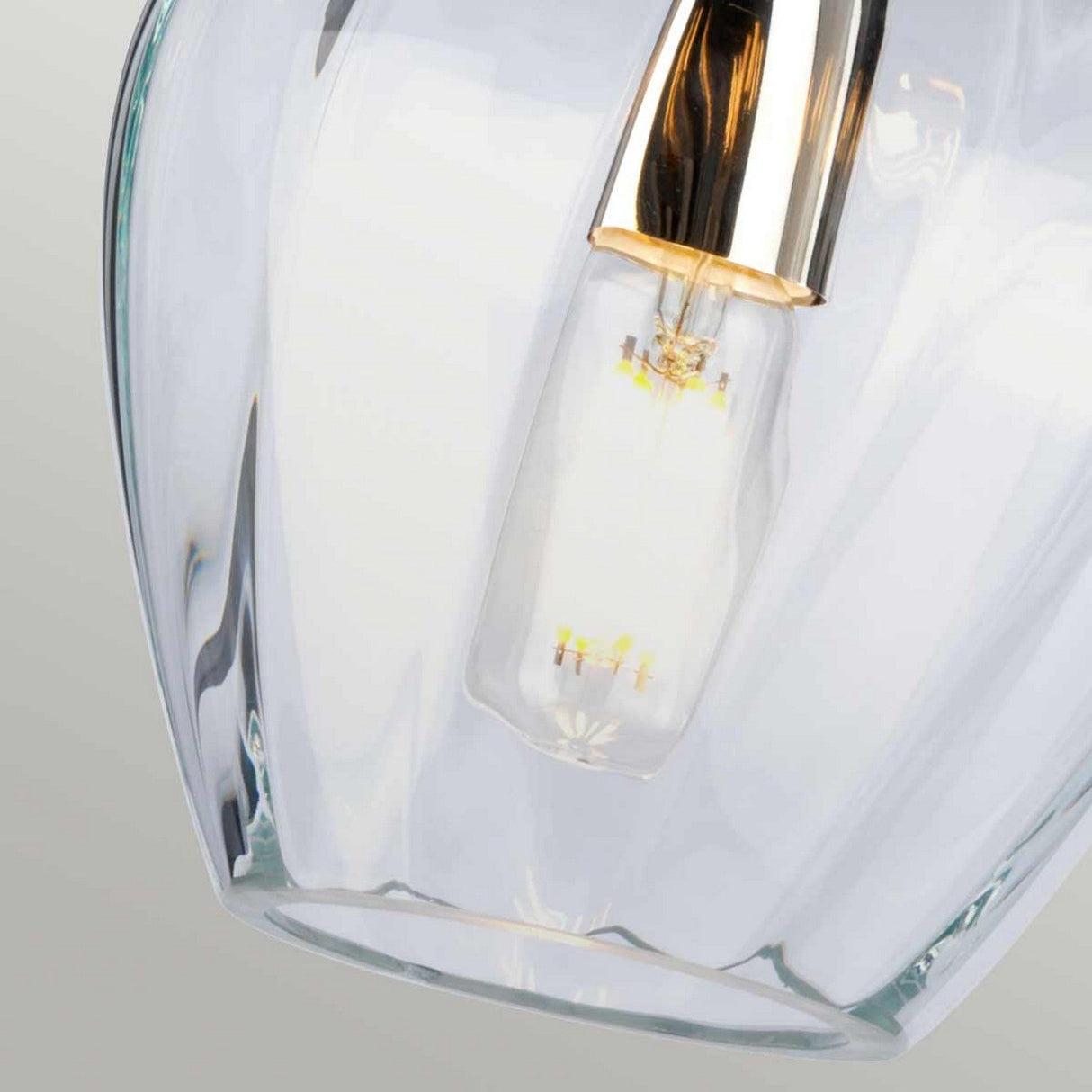 A close-up of the Tiber 1 Light Pendant - Polished Nickel/Clear Glass showcases its clear glass shade and visible bulb, topped with polished nickel. The illuminated bulb emits a soft glow against a neutral gray background.