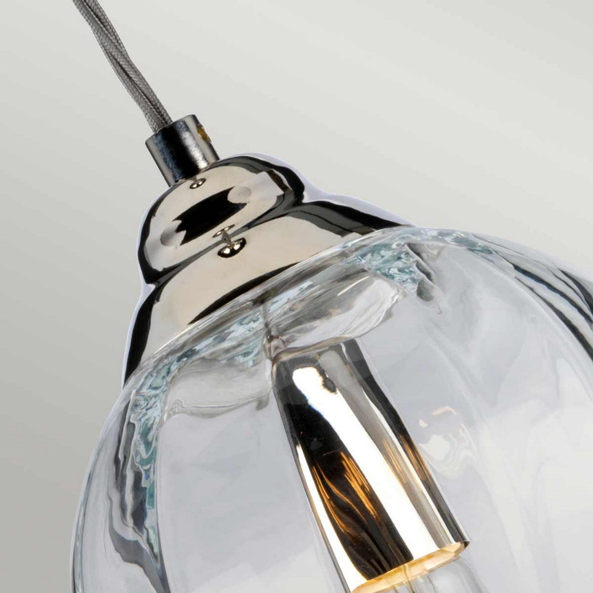 Close-up of the Tiber 1 Light Pendant featuring a clear glass shade and a polished nickel fixture on top. The illuminated bulb casts a warm glow, accentuating the sleek curves of the transparent glass against a soft gray background.