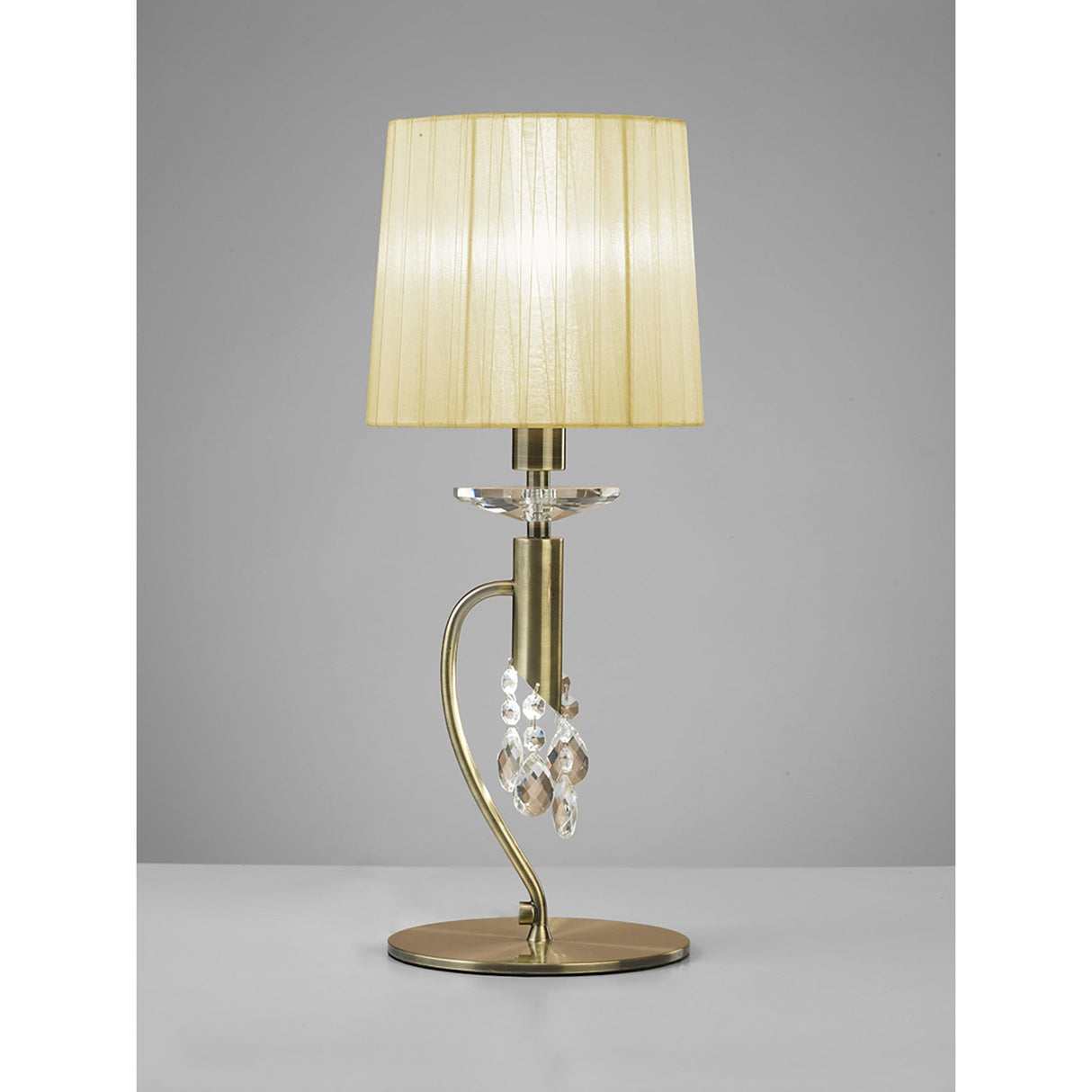The Tiffany 2 Light Crystal Table Lamp features a brass finish, cream shade, hanging crystal accents, and a curved metal base. It is displayed on a light gray surface against a neutral gray background.