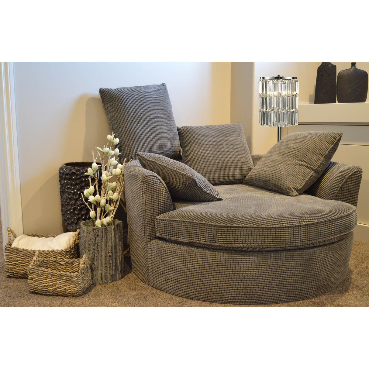 A cozy gray circular armchair with three cushions sits in a corner. Nearby, decorative baskets, a textured vase with white flowers, and the Torre 3 Light Crystal Curtain Table Lamp in polished chrome rest on a beige carpet.