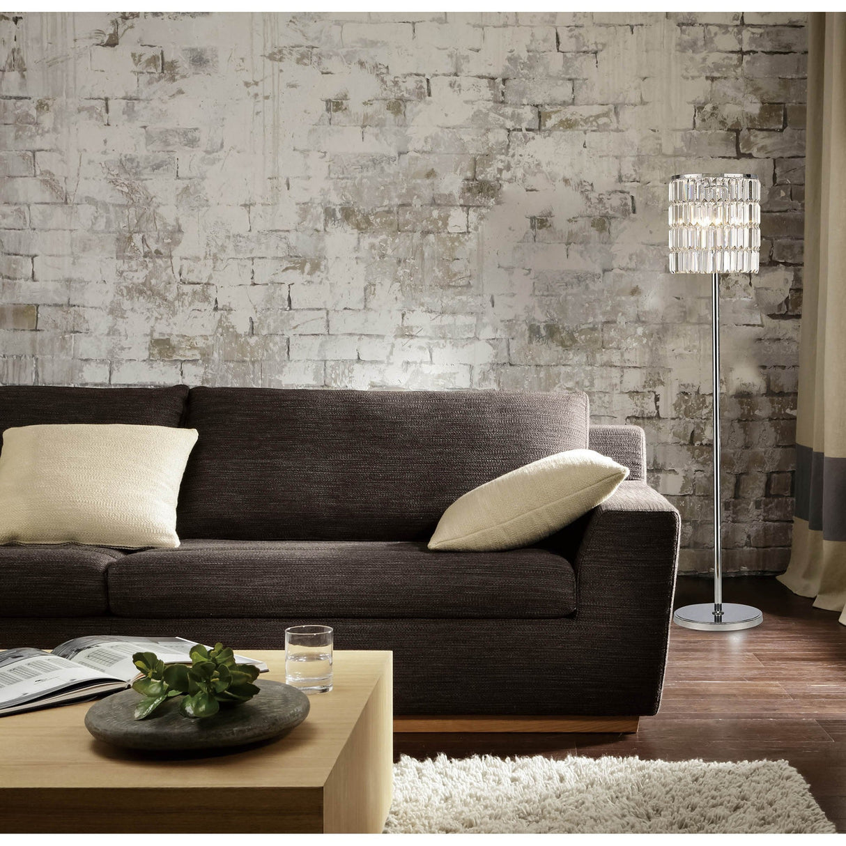 A modern living room features a dark brown sofa, beige cushions, a wooden coffee table with a plant, book, and glass of water. The Torre 5 Light Crystal Curtain Floor Lamp in polished chrome adds statement lighting to the gray and white brick wall.