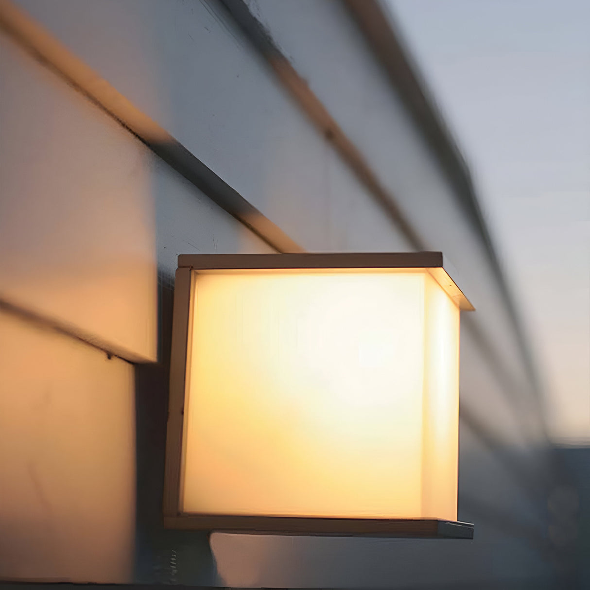 The Torsten 1 Light Outdoor Wall Fitting in graphite, featuring a square design, is mounted on a light-colored exterior wall. It emits a soft glow, highlighting its weather-resistant quality as the out-of-focus horizon suggests either dawn or dusk.