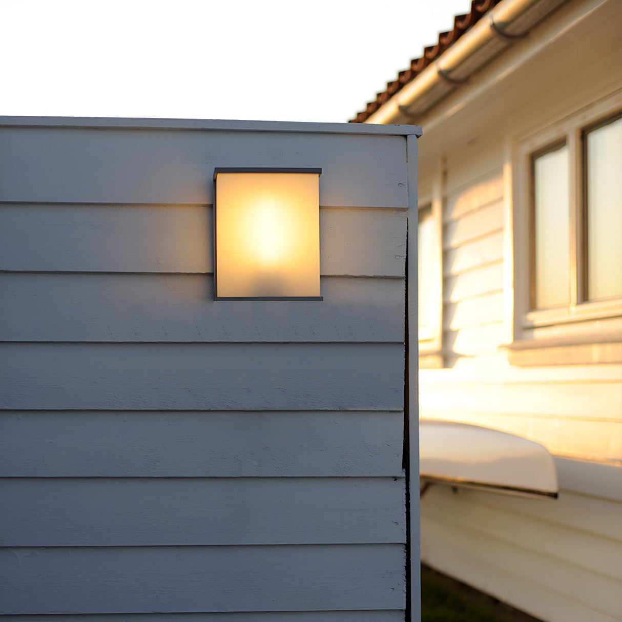 The Torsten 1 Light Outdoor Large Wall Light in a graphite finish casts a soft, warm glow on a white wooden wall during sunset, with the minimalist design highlighting the horizontal siding and a white house visible in the background.