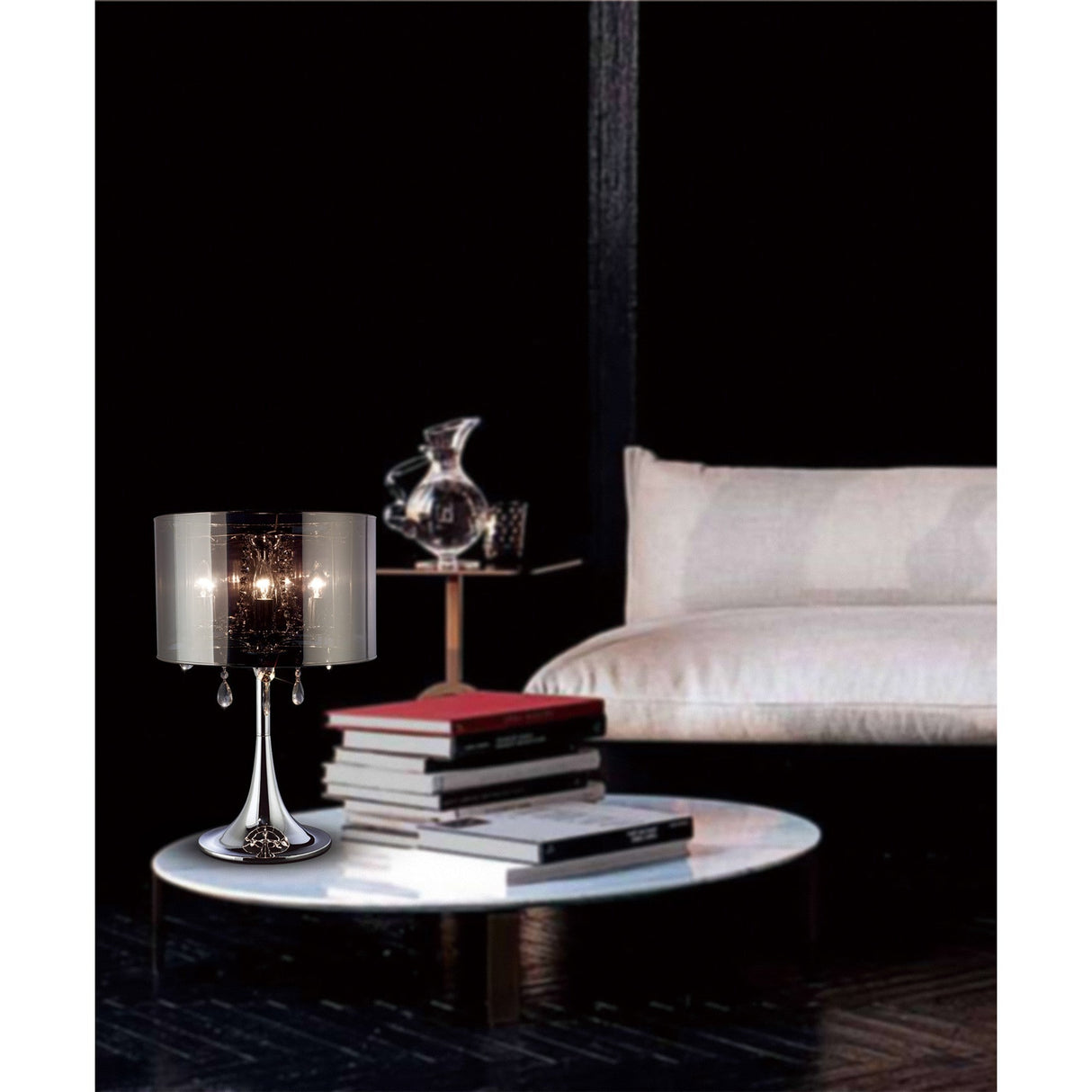 A modern living room with a white sofa, round white coffee table with books, glass decanter, and the Trace 3 Light Crystal Table Lamp in chrome with a chrome shade, all set against a dark wall.