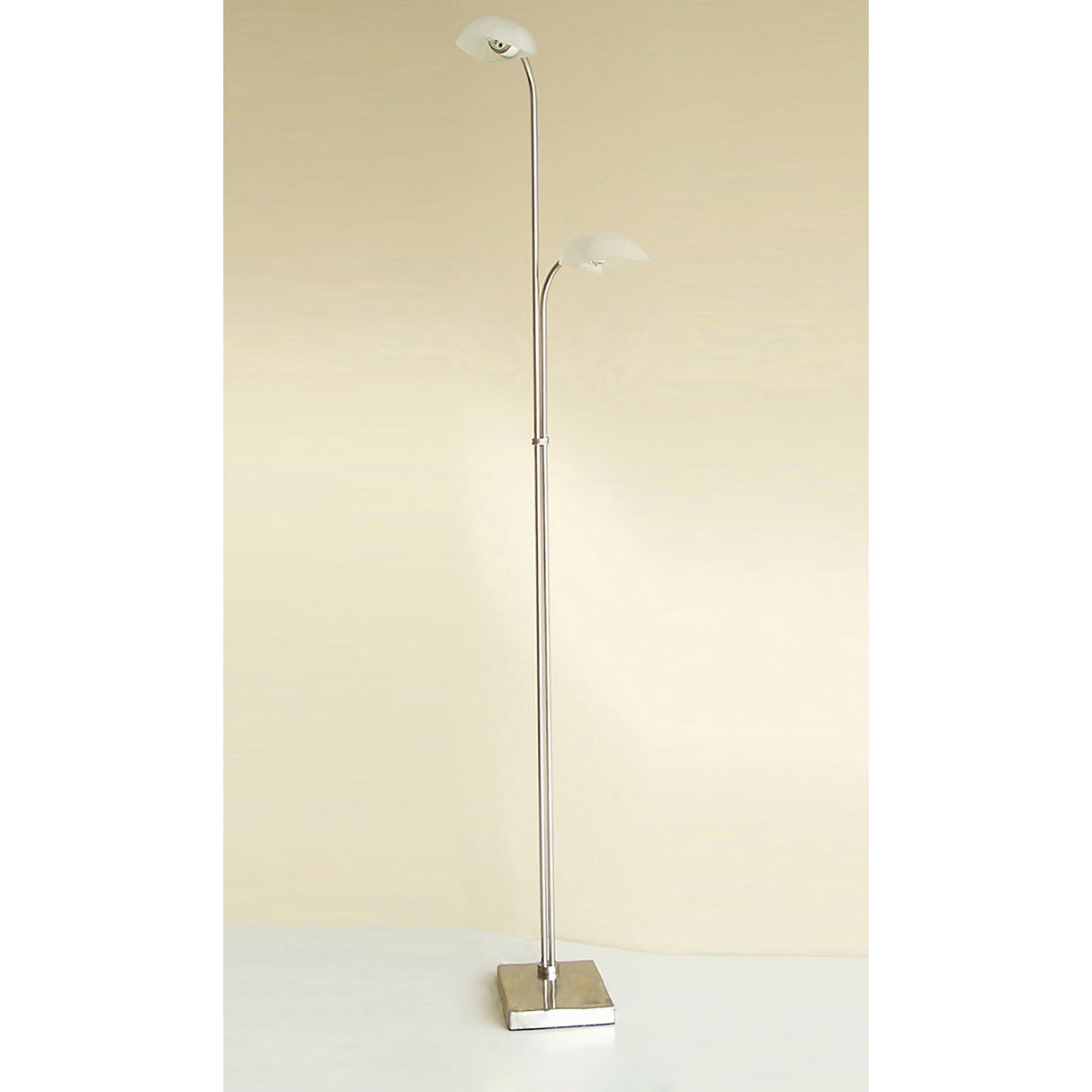 The Udine 2 Light Frosted Glass Floor Lamp in satin chrome features a square metal base, dual slender poles, and upward-facing frosted glass shades set at staggered heights against a plain beige background.
