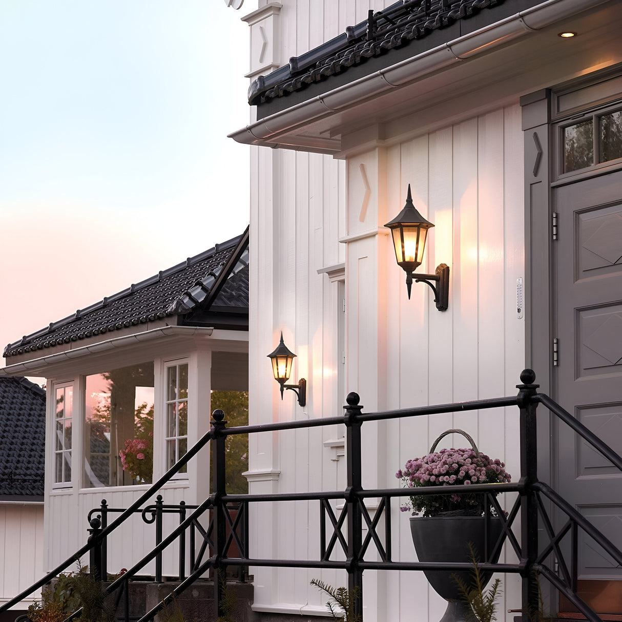 Front view of a white house featuring a striking black roof and decorative Valencia Outdoor 1 Light Up Lanterns—crafted from die-cast aluminum. A potted plant with pink flowers sits on a railing near the entrance, set against a pinkish sky that suggests early morning or evening, enhancing the glow of the lantern.
