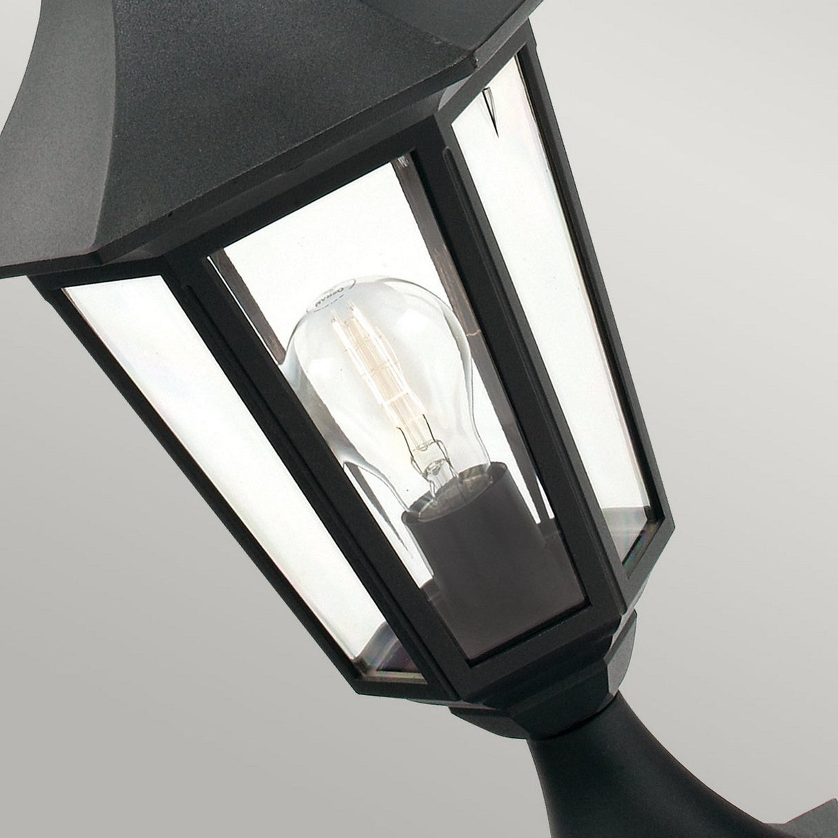 Close-up of the Valencia 1 Light Pedestal - Black, an outdoor lantern crafted from die-cast aluminum with clear glass panels, showcasing an exposed light bulb inside. The traditional, angular design of this outdoor lighting piece stands out against a neutral gray background.