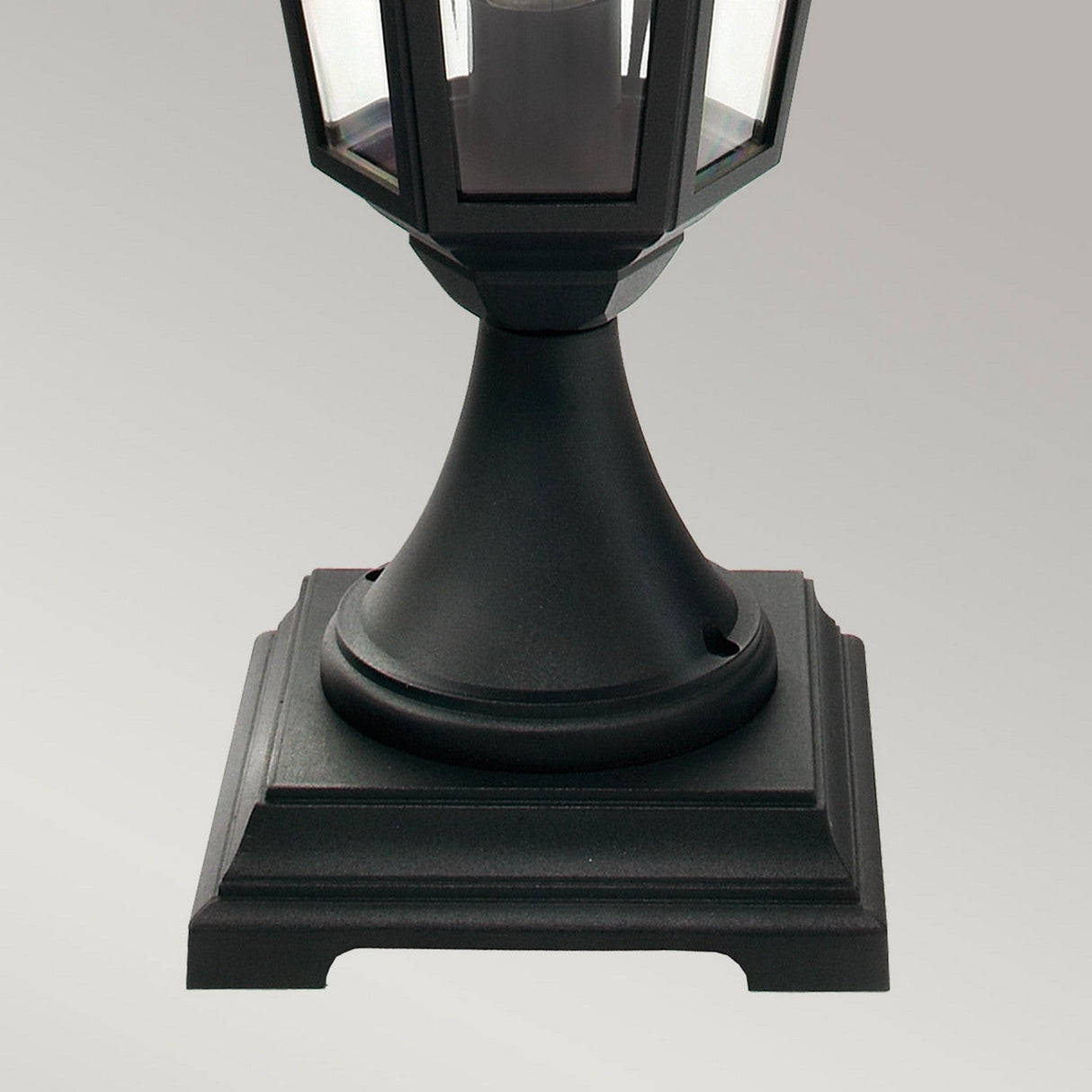 A close-up of the Valencia 1 Light Pedestal - Black highlights its black, hexagonal lantern design made from die-cast aluminum and resting on a square base. Positioned against a plain gray background, it features clear glass panels for a classic look.