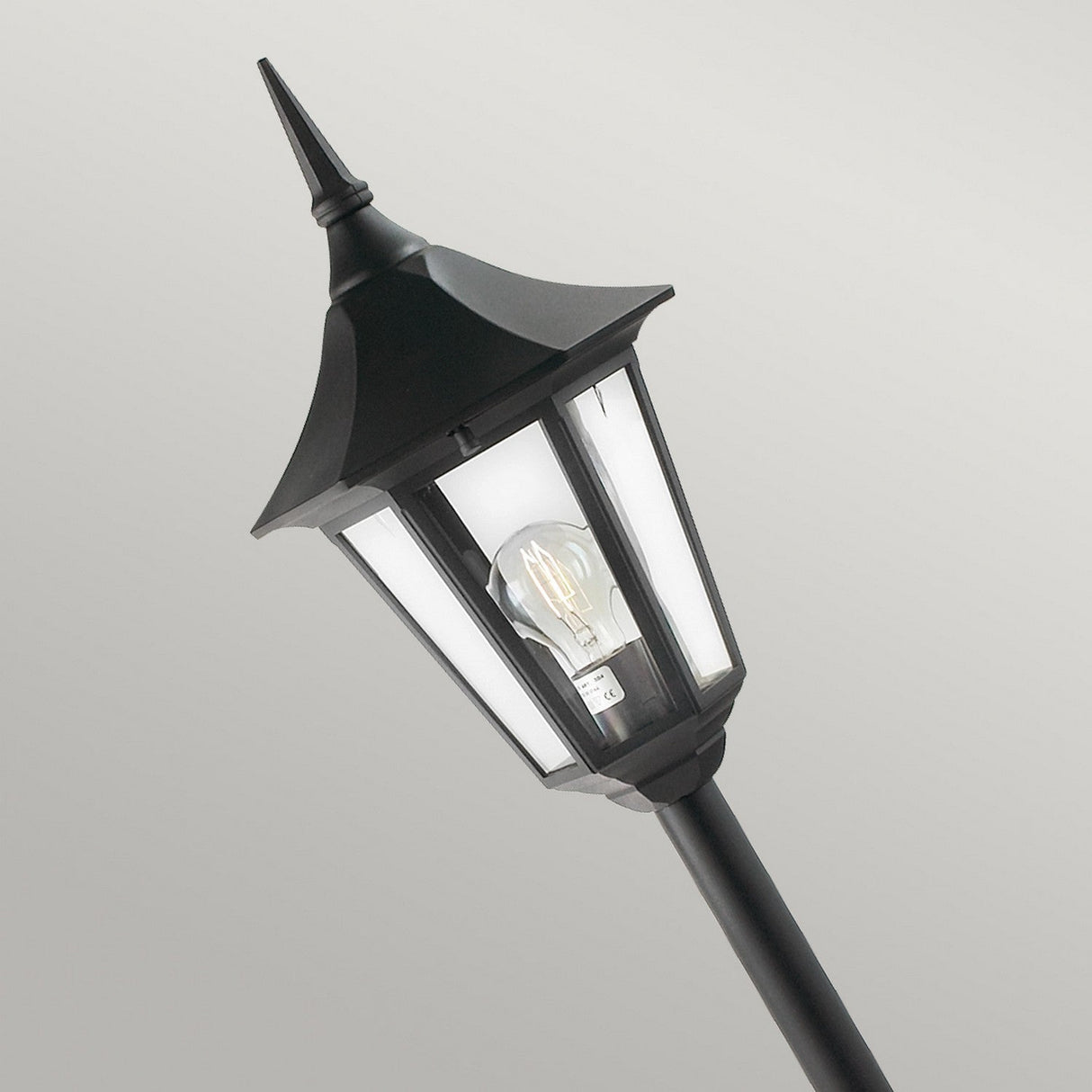 A black street lamp in a vintage style, featuring frosted glass panels and a visible light bulb, reminiscent of the elegant Valencia 1 Light Pillar - Black. This outdoor fixture is slightly tilted against a neutral gray background, making it ideal for illuminating garden pathways.