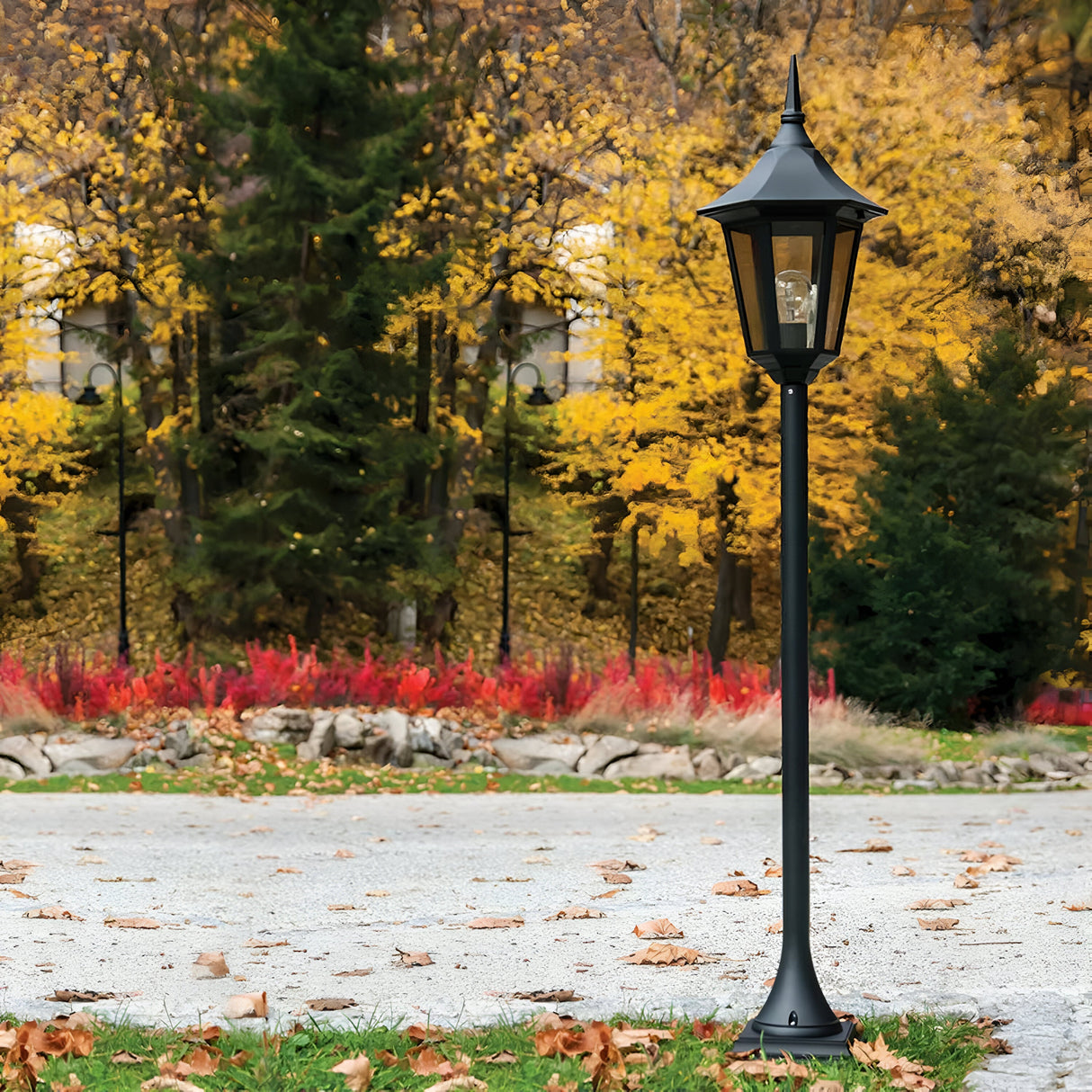 A Valencia 1 Light Pillar - Black, an elegant outdoor fixture, stands on a grassy area amidst fallen autumn leaves. Behind it, tall trees with vibrant yellow foliage and red bushes form a vivid natural backdrop, perfect for illuminating garden pathways.
